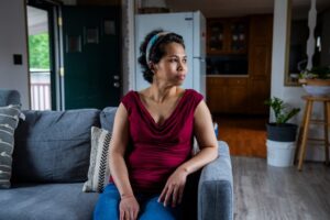 Jennifer Le, a woman in a red blouse and jeans, is sitting on her couch at home.